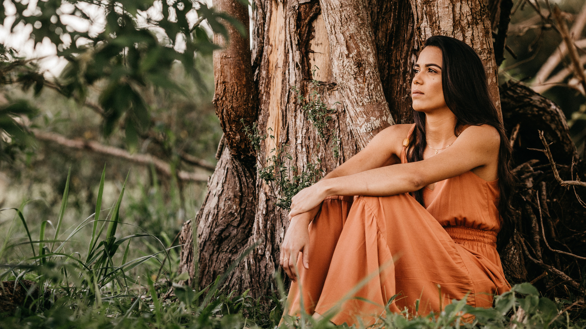 woman in orange dress sits near tree and reflects in nature